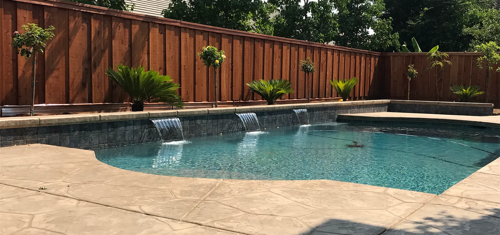 A V.O. pool: three sheer waterfalls pour from a dark tile wall into turquoise water, with a stamped concrete deck and a cedar fence behind.