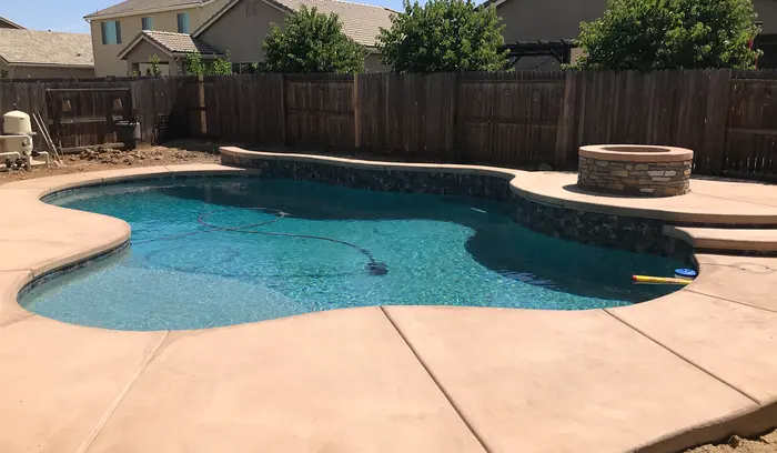 A free-form pool with a stacked-stone fire pit on a raised deck at the far end and a tan concrete deck in front.