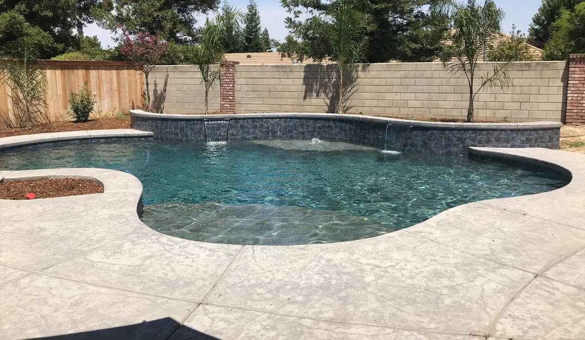A free-form pool with a raised wall of blue-gray tile, two sheer waterfalls, a bubbler on the shallow ledge and a planter cut into the deck.