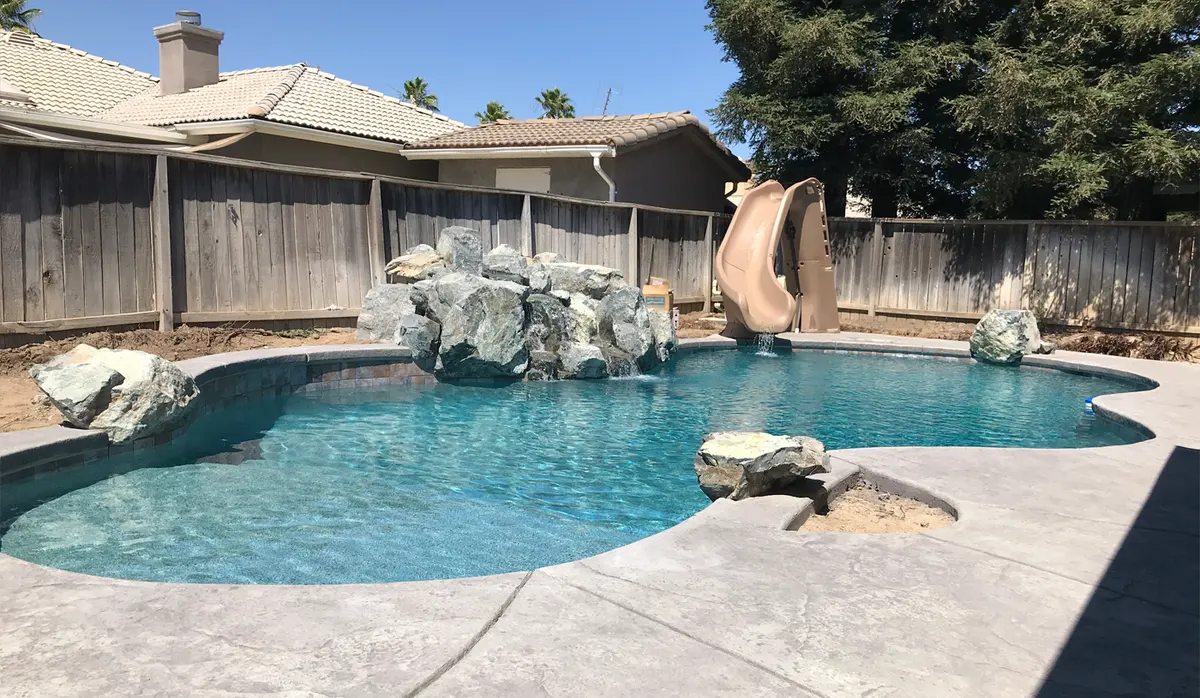 A free-form pool with a waterfall of large boulders and a tan slide, the new deck poured and the yard behind still bare dirt.
