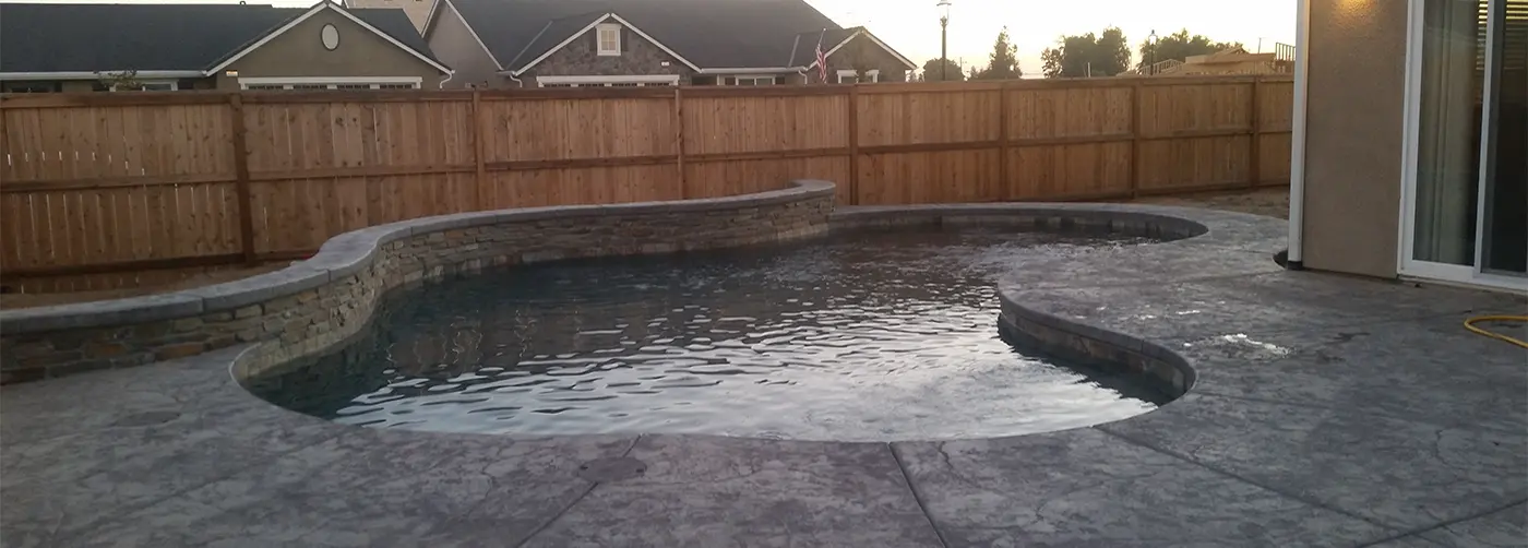 A free-form pool at dusk with a curved raised wall of stacked stone and a gray stamped concrete deck.